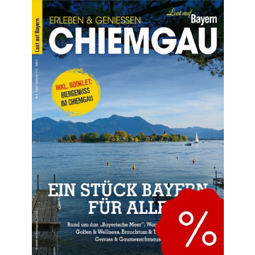 Blick auf einen See mit Segelboot, Steg und bewaldeter Insel vor Bergkulisse unter blauem Himmel.