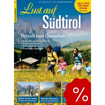 Familie mit zwei Kindern wandert durch blühende Wiese vor Berghütte und grünen Hügeln im Hintergrund.