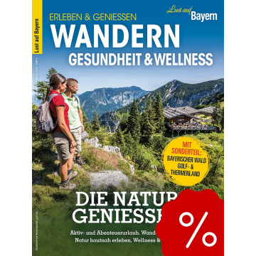 Zwei Wanderer mit Rucksack stehen auf einem Bergpfad und blicken auf bewaldete Berge und eine Hütte.