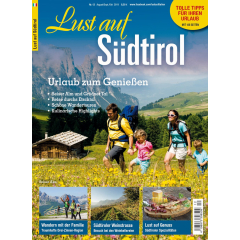 Familie mit zwei Kindern wandert durch blühende Wiese vor Berghütte und grünen Hügeln im Hintergrund.